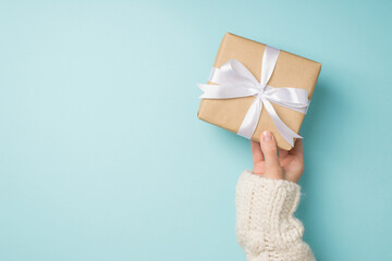 First person top view photo of hand in white knitted pullover holding craft paper giftbox with white satin ribbon bow on isolated pastel blue background with empty space