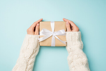 First person top view photo of hands in white knitted sweater holding craft paper giftbox with...