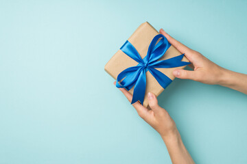 First person top view photo of hands giving craft paper giftbox with vivid blue ribbon bow on isolated pastel blue background with empty space