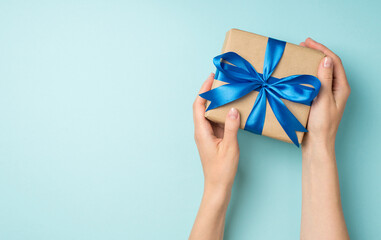 First person top view photo of female hands holding craft paper giftbox with blue ribbon bow on isolated pastel blue background with copyspace