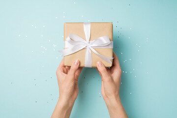 First person top view photo of hands holding craft paper giftbox with white ribbon bow over sequins on isolated pastel blue background