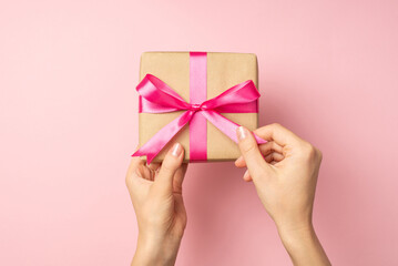 First person top view photo of hands unpacking craft paper giftbox with pink ribbon bow on isolated pastel pink background