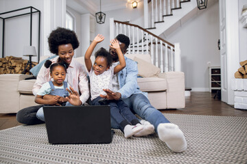 Two pretty girls and their young parents playing video games on modern laptop while sitting on floor. African american family using technology for entertainment at home.