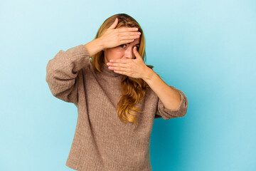 Caucasian woman isolated on blue background blink at the camera through fingers, embarrassed covering face.