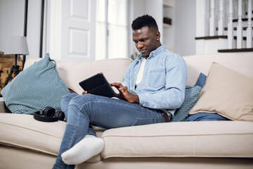 African american man surfing internet or making shopping online on digital tablet while sitting on cozy sofa. Young guy in casual outfit relaxing at home with modern gadget in hands.