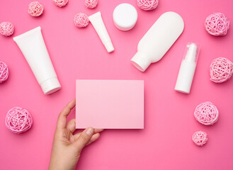 Female hand holding empty pink paper on a pink background and jar, bottle and empty white plastic tubes for cosmetics on a pink background