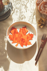Orange maple leaf decoration on plate. Autumn table setting with sunlight and cutlery. Top view