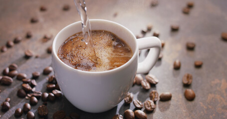 Pouring hot water into the cup of coffee and roasted coffee beans near the cup on the wooden table. Top view.