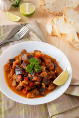 Hot eggplant stew decorated with lime on plate for healthy dinner at home. Homemade vegan Aubergine saute. Braised vegetables in a white bowl on wooden table.
