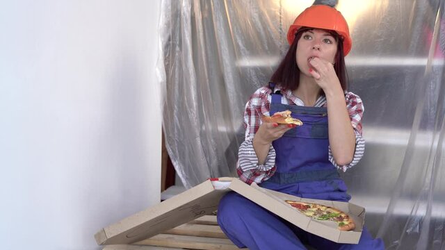 Pretty young woman builder with helmet working on apartment renovation. Construction, repair and renovation. took a break and is eating pizza.