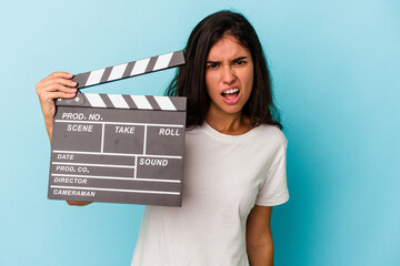 Young caucasian woman holding a clapperboard isolated on blue background screaming very angry and...
