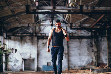 Fototapeta na wymiar Strong Muscular Handsome Man in Black Tank Top with Baseball Bat Walks in Empty Grunge Hall