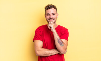 young handsome hispanic man looking happy and smiling with hand on chin, wondering or asking a...