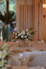 White tablecloths with clear vases and white flowers and fern arrangements. Golden colored plates, peavh napkins, table numbers and mirror centerpieces.