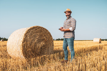 Naklejka na ściany i meble Happy farmer is standing beside bales of hay. He is examining straw after successful harvesting.