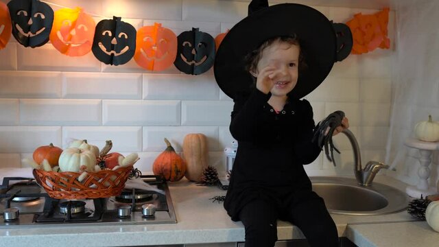 Child girl sitting on the kitchen table In Halloween Costume and playing with decorations. Halloween party at home