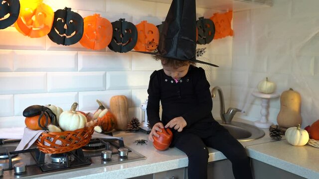 Child girl in halloween costume witch decorating pumpking with false eye lahes sitiing on kitchen table with garland and spiders