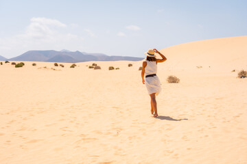 Fototapeta na wymiar A young Caucasian tourist in a white dress and a hat walking through the dunes of the Corralejo Natural Park, Fuerteventura, Canary Islands. Spain