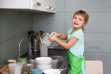 A little cute child girl washing dishes in the white kitchen interior. casual lifestyle photo series in real life interior. Child with helping his parents with housework.
