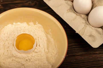 A broken white chicken egg in a bowl of wheat flour and several whole eggs in a tray made of white cardboard, close-up selective focus.