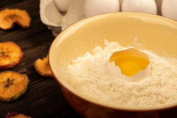 A broken white chicken egg in a bowl with wheat flour, dried apple chips and several whole eggs in a tray made of white cardboard, close-up selective focus.