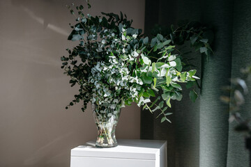 White table and glass vase with eucalyptus twigs against white wall in hight contrast. Mininmalistic composition with deep shadows