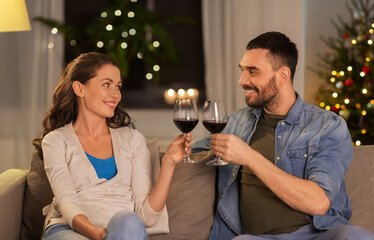 leisure, clebration and winter holidays concept - happy couple drinking red wine at home in evening over christmas tree lights on background