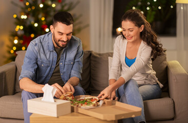 winter holidays, people and fast food concept - happy couple eating takeaway pizza at home in evening over christmas tree lights background