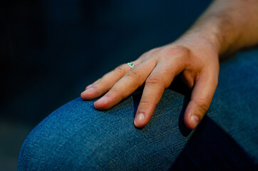 a man's elegant hand with an engagement ring lies calmly on his knee in jeans