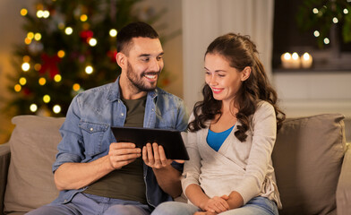 technology, winter holidays and people concept - happy couple using tablet computer at home in evening over christmas tree lights on background