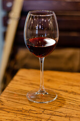 an elegant transparent tall glass with red wine stands on a light brown wooden table and lights are reflected in it