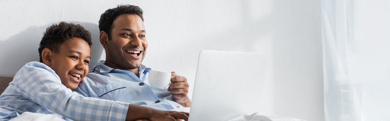 laughing african american boy pointing at laptop while watching movie with dad in bedroom, banner