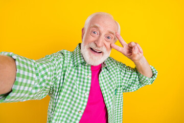 Self-portrait of attractive cheerful funky grey-haired man showing v-sign having fun isolated over bright yellow color background