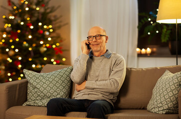 technology, winter holidays and communication concept - happy smiling senior man calling on smartphone at home in evening over christmas tree lights on background