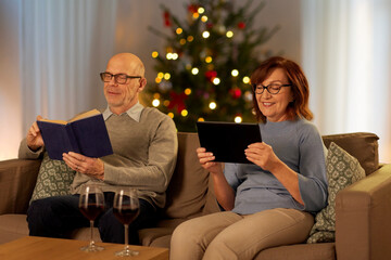 leisure and winter holidays concept - happy senior couple reading book and using tablet pc computer at home in evening over christmas tree lights on background