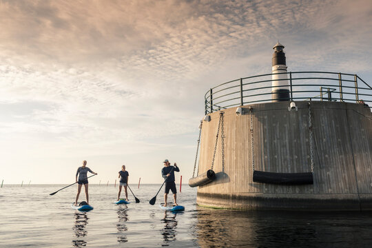 Female friends rowing paddleboard with male instructor by lighthouse in sea