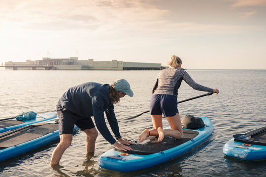 Woman learning paddleboarding from male instructor in sea during sunset