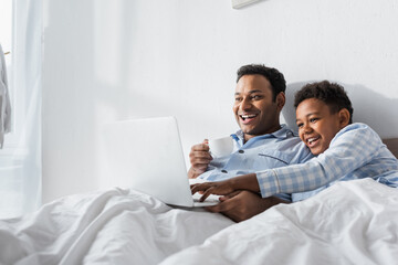 Fototapeta premium excited african american boy and dad watching comedy on laptop in morning in bed