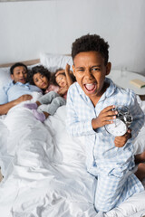 Fototapeta na wymiar thrilled african american boy with alarm clock shouting near blurred family lying in bed