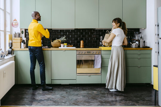 Full Length Of Smiling Couple Talking With Each Other While Doing Chores In Kitchen At Home