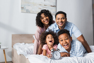 excited couple near kids and alarm clock smiling at camera in bedroom