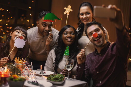 holidays, people and celebration concept - multiethnic group of happy friends with party props having christmas dinner at home and taking selfie with smartphone