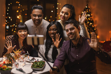 holidays, party and celebration concept - multiethnic group of happy friends having christmas dinner at home and taking selfie with smartphone and waving hands