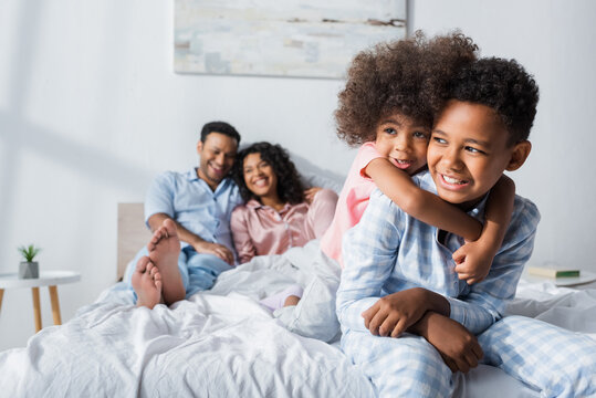 cheerful african american girl hugging brother while having fun near blurred parents in bedroom