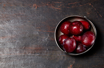 Ripe red plums in bowl with water drops. Juicy fruit on wooden background, closeup. Healthy food on dark kitchen table