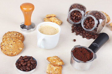Cup of espresso, crackers, cookies, holder with ground coffee, tamper and cans of coffee beans on table.