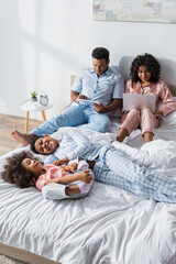 african american couple using gadgets near kids having fun on bed