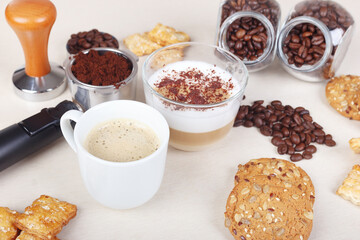 Cup of espresso, cappuccino with chocolate crumbs, cookies, crackers, holder with ground coffee, tamper and coffee beans on table.