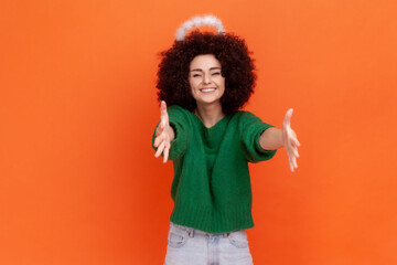 Come to my arms! Smiling angelic woman with Afro hairstyle in green casual style sweater and nimb over head outstretching hands, wants to embrace you. Indoor studio shot isolated on orange background.