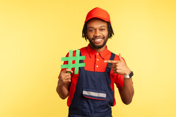 Portrait of happy workman wearing blue overalls standing, holding green hashtag and pointing finger to it, looking at camera with smile. Indoor studio shot isolated on yellow background.